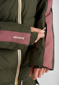 En närbild av en persons hand som justerar en dragkedja på ärmen av en grön jacka med rosa detaljer och en logotyp som säger "BURTON."