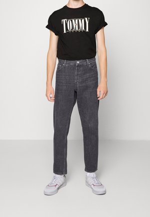 Osoba oblečená v černém tričku "Tommy Jeans", tmavě šedých džínech s vysokým pasem a bílých teniskách stojící proti světle šedému pozadí.