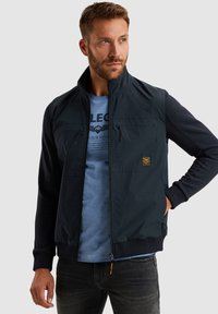 Giacca zip navy con colletto alto, tessuto leggero, maniche navy a contrasto, due tasche frontali e toppa con logo sul petto.