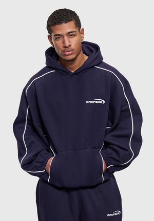 Jeune homme aux cheveux courts et bouclés portant un sweat à capuche Dropsize bleu marine et un pantalon assorti avec des coutures blanches, les mains dans les poches du sweat, fond neutre.