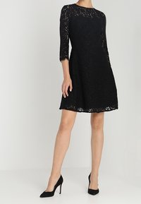 Robe en dentelle noire avec manches trois-quarts, col rond et silhouette trapèze. Portée avec des chaussures à talons hauts noires.