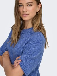 Stickad blå tröja med korta ärmar, rund halsringning och ett texturerat mönster. Modellen står med armarna i kors och bär små ringörhängen.