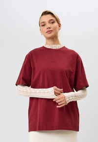 Burgundska bombažna majica s kratkimi rokavi, preložena čez dolgo belo čipkasto bluzo, z okroglo izrezom in sproščenim krojem.