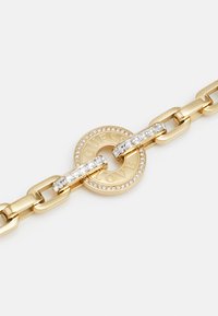 Guldfarvet metalarmbånd med en rund charm, der er gravert med "GUESS LOVE," prydet med klare sten og sammenflettede kædelinker.