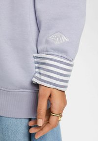 Sudadera de color púrpura claro con una tela texturizada, dobladillo acanalado y puños a rayas con un emblema blanco en la manga.