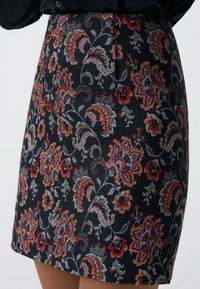 Jupe à motif floral en tissu foncé avec des teintes vives de rouge, rose et vert. Fini texturé avec un broderie détaillée et une coupe ajustée.