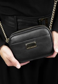 Zwarte gestructureerde leren schoudertas met gouden hardware en een rechthoekig plaatje, voorzien van een ritssluiting en een kettingstrap.
