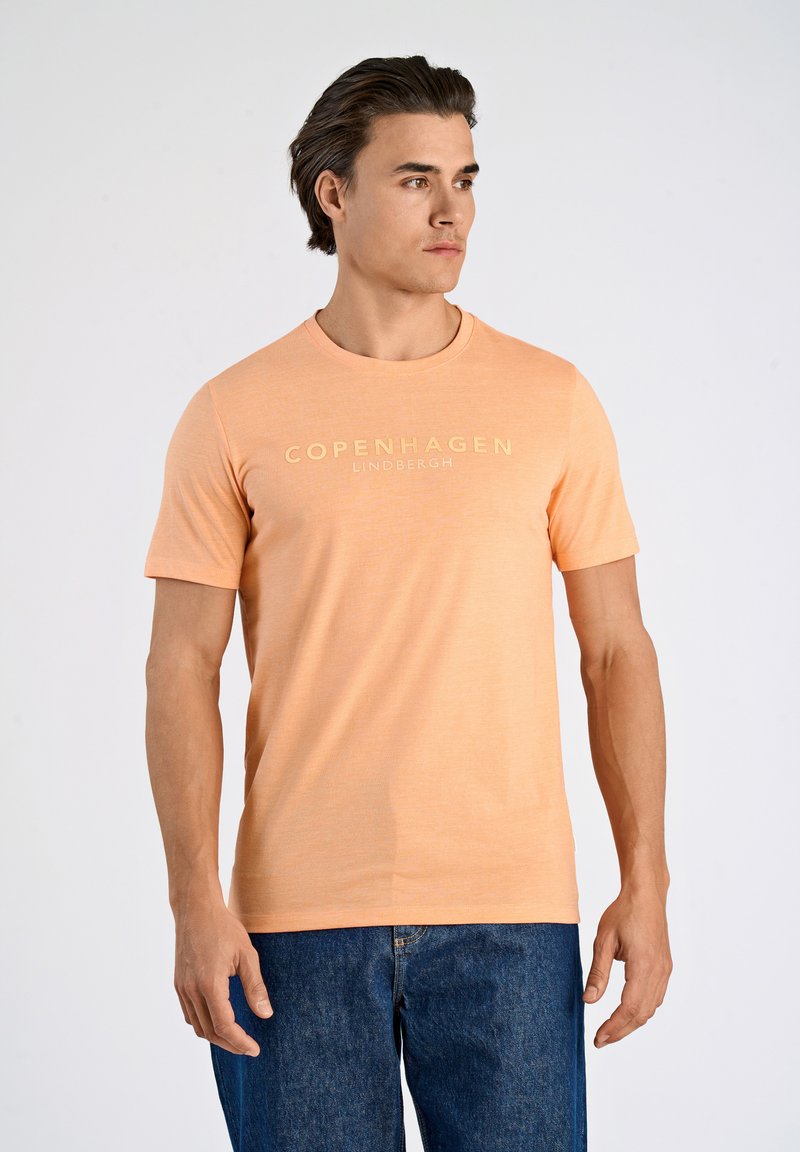 Camiseta de algodón de manga corta en color naranja claro que presenta "COPENHAGUE" y "LINDBERGH" estampados en texto color crema en el pecho.