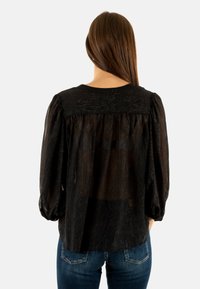 Blusa negra con espalda transparente, mangas largas abullonadas y una tela texturizada con sutiles hilos metálicos, combinada con jeans azules.