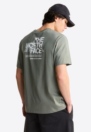 Junger Mann trägt ein grünes The North Face T-Shirt mit Berggrafik und dem Slogan "Never Stop Exploring", schwarze Shorts und eine schwarze Kappe.