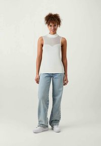 Top branco sem mangas com uma parte superior texturizada e canelada e uma parte inferior lisa, combinado com jeans azul claro e ténis brancos.
