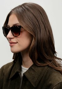 Occhiali da sole tartarugati con design angolare e lenti scure, abbinati a una giacca in camoscio verde scuro. I capelli sono acconciati in morbide onde.