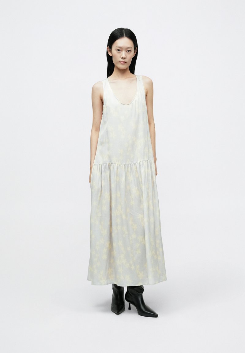 Lang, ærmeløs kjole i let stof med et subtilt gult blomstermønster, der har en rund halsudskæring og en indsnævret talje, kombineret med sorte støvler.