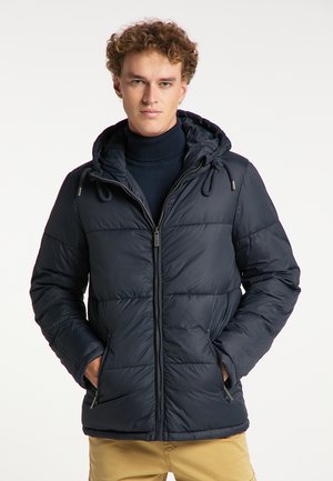 Jeune homme aux cheveux bouclés portant une doudoune bleu marine avec capuche et un pantalon beige, les mains dans les poches, regardant devant lui.