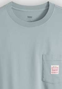 Ανοιχτό μπλε άνετο μπλουζάκι Levi's με στρογγυλή λαιμόκοψη, τσέπη στο στήθος και μια μικρή ετικέτα στην τσέπη με την ένδειξη "εργασία που πρέπει να γίνει."