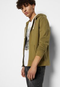 Oliven grønn hettegenser med glidelås, snor, frontlommer og ribbestrikkede mansjetter, båret over en hvit grafisk t-skjorte og svarte jeans.