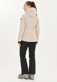 Veste isolante beige avec une capuche, associée à un pantalon noir et des bottes grises. Présente un design ajusté et des fermetures éclair aux poignets du pantalon.