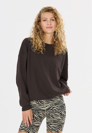 Kvinde med krøllet blondt hår iført en løs mørk bluse med lange ærmer og tætsiddende zebra-mønstrede shorts mod en ensfarvet hvid baggrund.