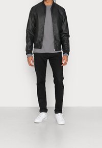 Man med svart läderjacka, grå långärmad tröja, svarta jeans och vita sneakers som står mot en enkel ljus bakgrund.