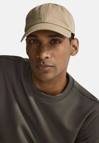 Beige bomullsbasketmössa med en böjd skärm, bärs av en person i en grå sweatshirt. Har sömmar på mössans krone och en slät yta.