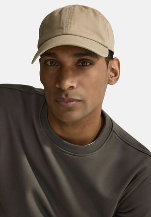 Beige bomuldsbaseballcap med en buet skygge, båret af en person i en grå sweatshirt. Har syninger på capens krone og en glat overflade.