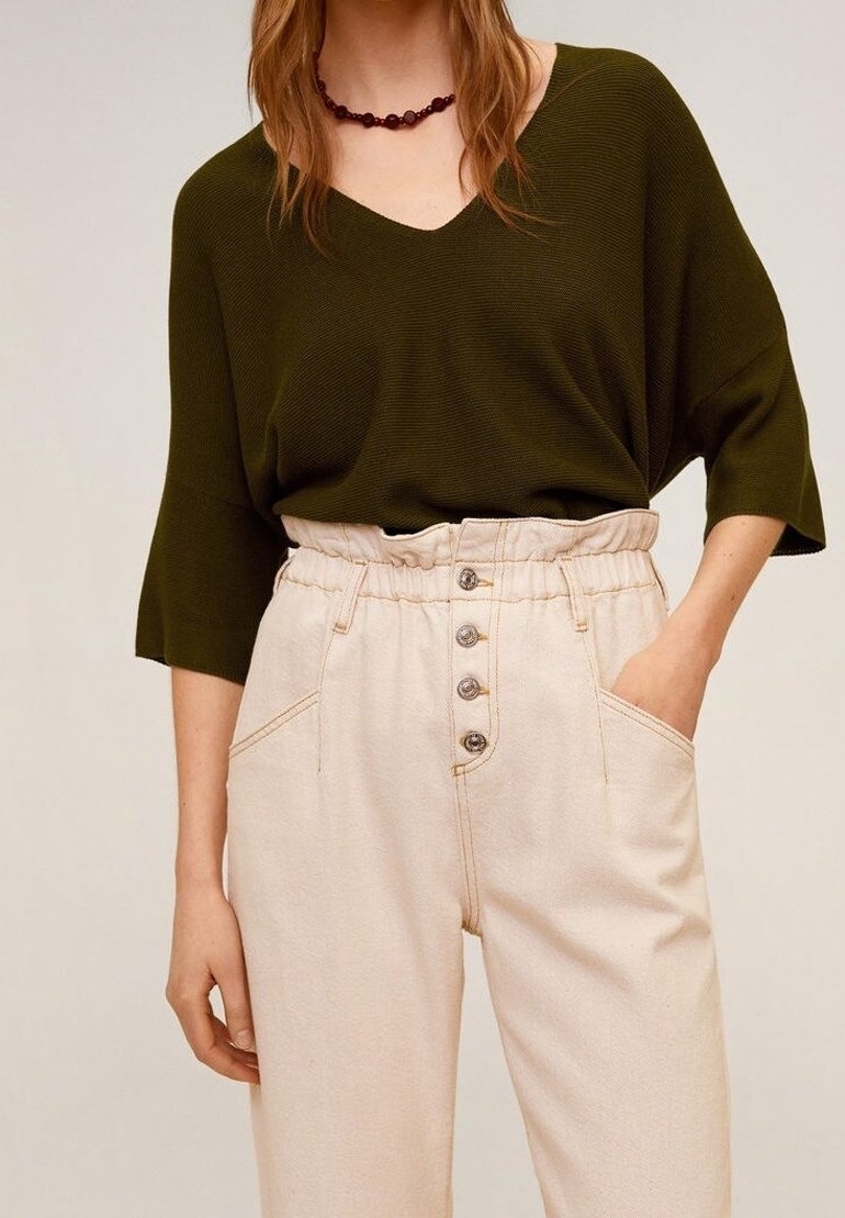 Personne portant un haut ample vert foncé à col en V et un pantalon beige taille haute avec braguette à boutons, une main dans la poche.