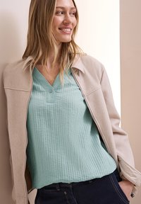 Giacca beige chiaro con zip sopra un top verde menta testurizzato con scollo a V e dettagli con bottone, abbinata a jeans di denim scuro. Contrasto di tessuti lisci.