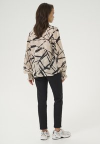 Lichte beige blouse met abstracte zwarte patronen, lange mouwen en gathered manchetten, gecombineerd met zwarte slim-fit broek en grijze sneakers.