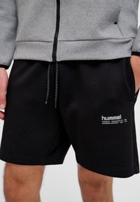 Férfi fekete Hummel rövidnadrágban, fehér logós felirattal és szürke cipzáras dzsekiben, kezek részben a zsebekben.