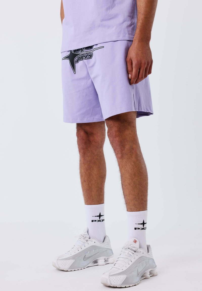 Homme portant un short violet clair avec un graphisme noir, des chaussettes blanches avec un logo noir, et des baskets blanches Nike sur un fond uni.