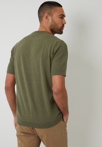 Olijfgroen gebreid sweater met korte mouwen en een gestructureerd patroon op de mouwen, gecombineerd met tan gekleurde broeken.