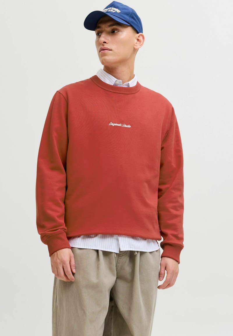 Czerwona bawełniana bluza z ribbowanymi ściągaczami i dekoltem, z białym haftowanym tekstem na piersi. Nałożona na paskowaną koszulę.