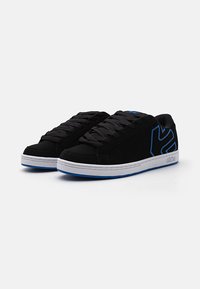 Chaussures de sport noires avec des accents bleus, en daim lisse, bout rond, semelle blanche plate et lacets tissés, avec un logo sur le côté.