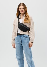 Černá crossbody taška s texturovaným povrchem, obdélníkovým tvarem, nastavitelným popruhem a minimalistickým designem, nošena přes béžovou bundu.