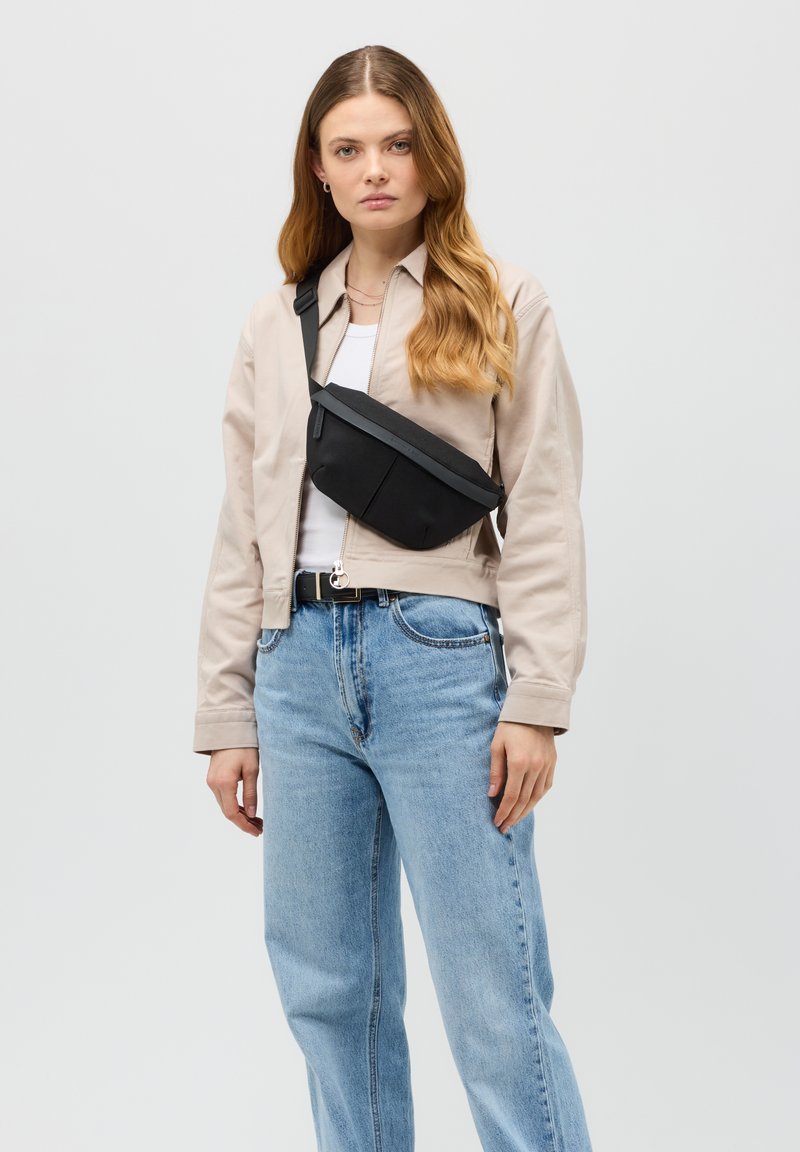 Černá crossbody taška s texturovaným povrchem, obdélníkovým tvarem, nastavitelným popruhem a minimalistickým designem, nošena přes béžovou bundu.