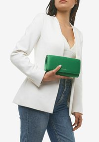 Clutch verde in finta pelle con una texture liscia e chiusura a zip, tenuto in mano, indossato con un blazer bianco e jeans in denim.