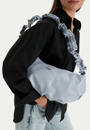 Žena v černém tričku a světle modrých džínách nese světle modrou kabelku s řaseným popruhem přes rameno.