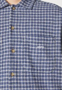 Chemise à carreaux bleue, blanche et noire boutonnée avec une poche poitrine arborant un petit logo blanc "obey".