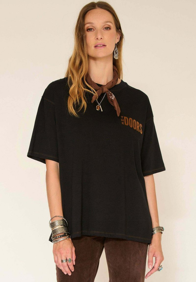 Femme aux longs cheveux ondulés portant un t-shirt noir surdimensionné "The Doors", une écharpe brune nouée autour du cou, des boucles d'oreilles en argent, plusieurs bracelets et bagues.