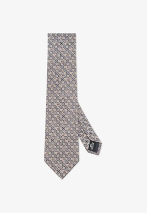 Wąski beżowy krawat z powtarzającym się niebieskim okrągłym wzorem. Posiada teksturowane sploty oraz małą czarną metkę na dolnym końcu.