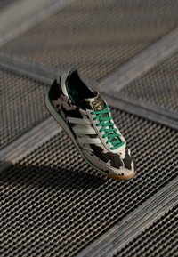 Cow-print sneaker featuring green laces, three white stripes, and a textured gum sole with a sturdy design and smooth surface.