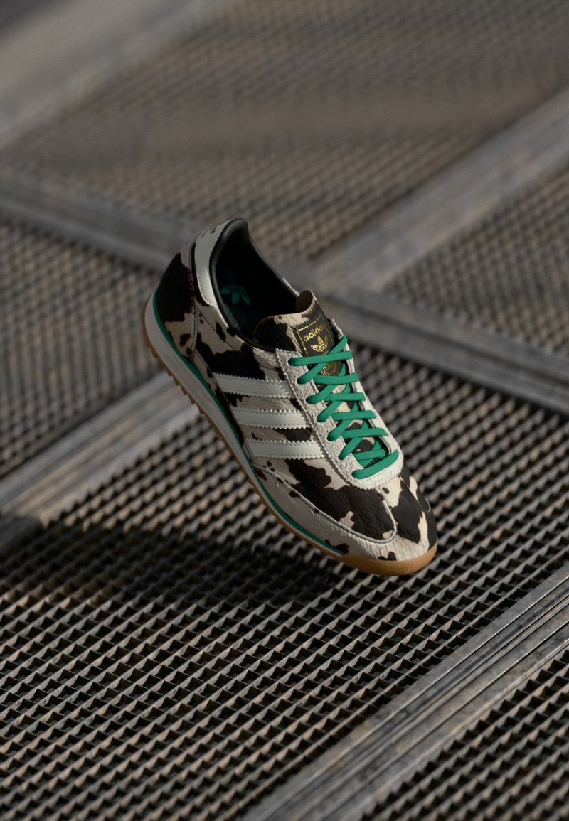 Cow-print sneaker featuring green laces, three white stripes, and a textured gum sole with a sturdy design and smooth surface.