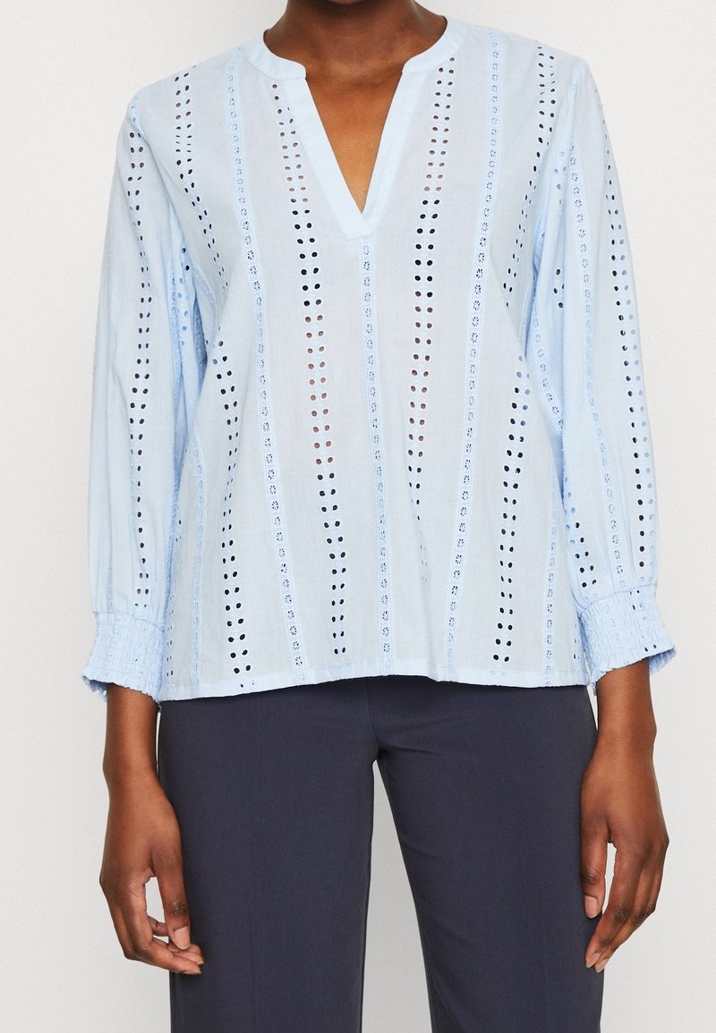 Blusa azzurro chiaro con ricami a strisce verticali e motivi ritagliati, dotata di scollatura a V e polsini elasticizzati, realizzata in tessuto traspirante.