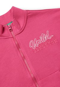 Sudadera rosa con cremallera hecha de tela suave que presenta "United Colors of Benetton" bordado en blanco. Incluye un cuello alto y un bolsillo delantero.