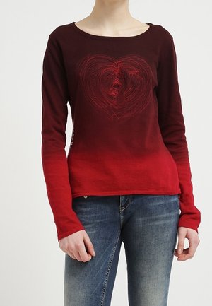 Mujer con una camiseta roja de manga larga con diseño de corazón en el pecho y vaqueros azules, de pie frente a un fondo liso.
