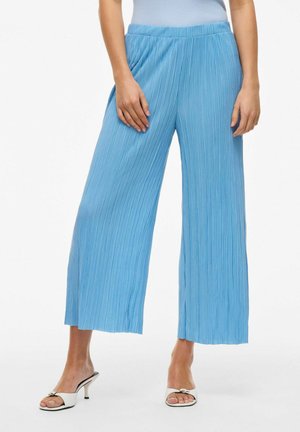 Pantalons large plissés bleu clair avec une taille élastique, présentant une texture lisse et une coupe ample, associés à des sandales à talons blanches.
