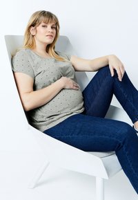 Camiseta gris con lunares y mangas cortas, combinada con jeans azul oscuro, usada por una persona embarazada sentada en una silla minimalista blanca.