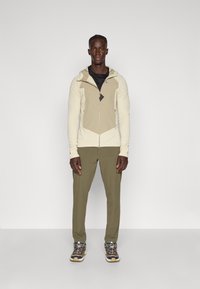 Casaco com capuz em creme e bege, com tecido texturizado, fecho de correr e detalhes em contraste; combinado com calças verde-azeitona e sapatos de exterior.