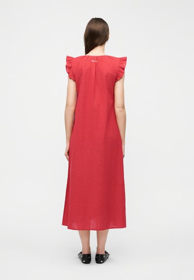 Γυναίκα με μακριά καστανά μαλλιά, φορώντας κόκκινο μίντι φόρεμα χωρίς μανίκια με λεπτομέρειες βολάν στους ώμους και μαύρα επίπεδα παπούτσια, που στέκεται με την πλάτη προς τα εμπρός.