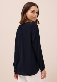 Blusa blu navy a maniche lunghe con colletto arricciato e vestibilità rilassata, caratterizzata da una texture liscia e un orlo curvo.